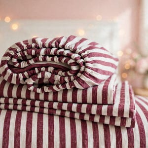 May include: A set of bed linens featuring a pattern of vertical stripes in deep burgundy and off-white. The collection includes a fitted sheet with elasticized edges and several folded flat sheets, all displaying the same striped design.