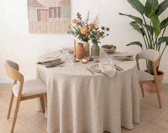 Könnte beinhalten: Ein runder Esstisch mit einer beigen Leinentischdecke, eingedeckt mit Tellern, Servietten und Besteck. Zwei Holzstühle mit beigen Polstersitzen stehen am Tisch, der mit einer zentralen Blumenvase und einer Kerze dekoriert ist.