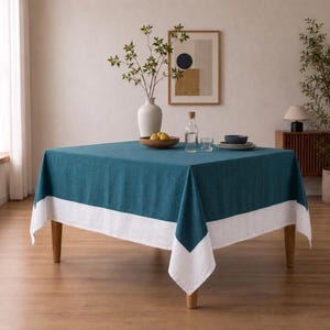 Puede incluir: Un mantel cuadrado que presenta un centro de color verde azulado intenso y un borde ancho de color blanco s&oacute;lido a lo largo del borde inferior. La tela tiene una textura tejida visible y cae sobre los cuatro lados de una mesa de madera, creando un aspecto bicolor.