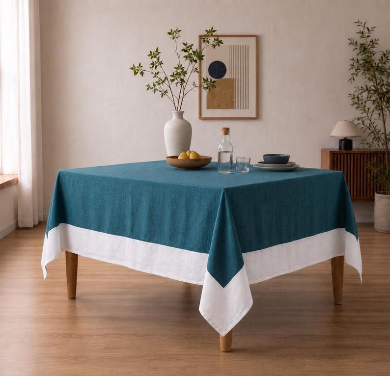 Puede incluir: Un mantel cuadrado que presenta un centro de color verde azulado intenso y un borde ancho de color blanco s&oacute;lido a lo largo del borde inferior. La tela tiene una textura tejida visible y cae sobre los cuatro lados de una mesa de madera, creando un aspecto bicolor.