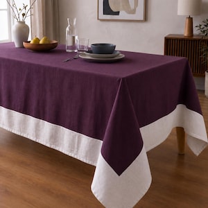 Puede incluir: Un mantel rectangular de color morado ciruela intenso que presenta un borde ancho y liso de color blanco roto a lo largo del borde inferior. La tela tiene una textura tejida visible y el mantel cuelga por los lados de una mesa de comedor de madera.