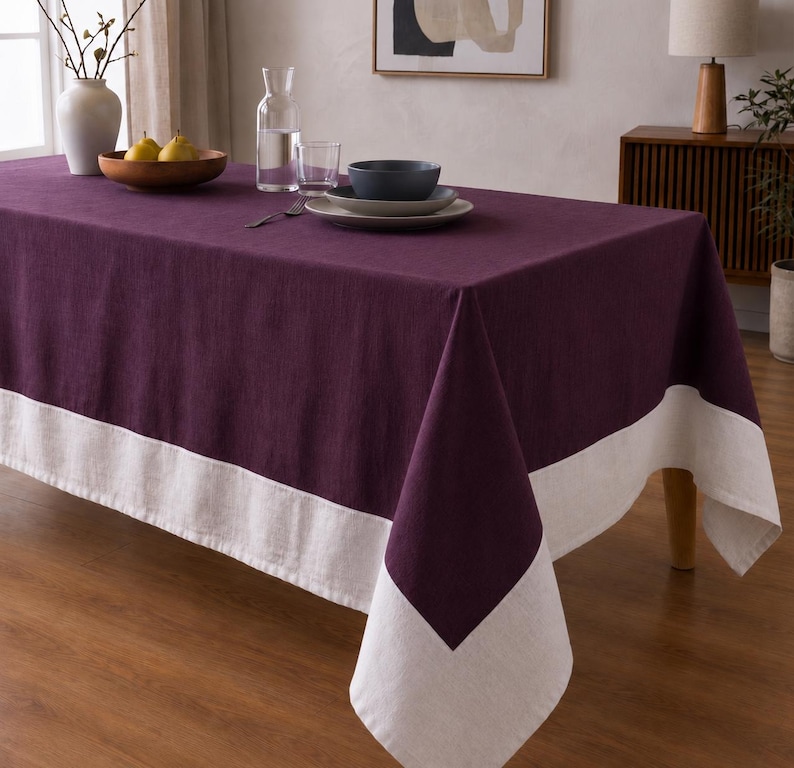 Puede incluir: Un mantel rectangular de color morado ciruela intenso que presenta un borde ancho y liso de color blanco roto a lo largo del borde inferior. La tela tiene una textura tejida visible y el mantel cuelga por los lados de una mesa de comedor de madera.