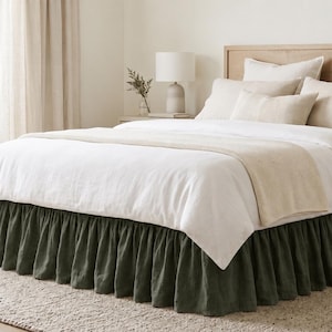 May include: A bed featuring a dark olive green ruffled bed skirt, a white duvet cover, and a light beige runner. The bed is styled with multiple pillows in shades of white and beige, set against a light wood headboard and frame.