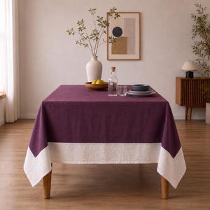 Puede incluir: Una mesa de comedor rectangular cubierta con un mantel de lino de color morado intenso que presenta un borde ancho y blanco en contraste en la parte inferior. La mesa est&aacute; decorada con un jarr&oacute;n blanco con ramas, una botella y cuencos.