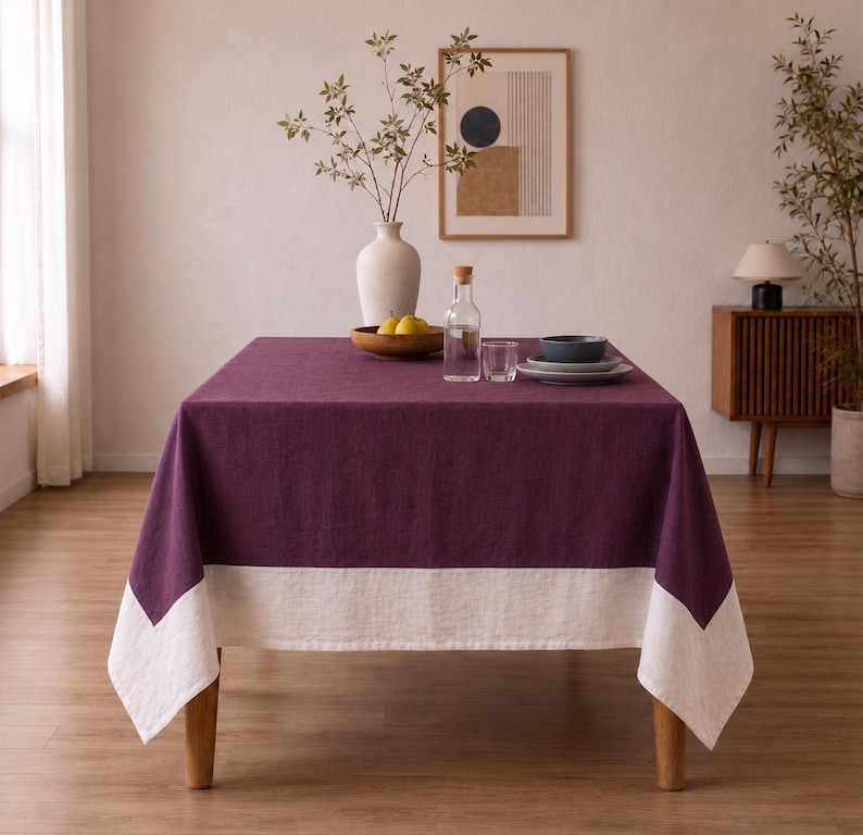 Puede incluir: Una mesa de comedor rectangular cubierta con un mantel de lino de color morado intenso que presenta un borde ancho y blanco en contraste en la parte inferior. La mesa est&aacute; decorada con un jarr&oacute;n blanco con ramas, una botella y cuencos.