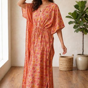 Puede incluir: Un vestido caft&aacute;n largo hasta los pies con un estampado floral en tonos coral y naranja, con cuello en V, mangas anchas estilo kimono y un cord&oacute;n ajustable en la cintura. La prenda tiene aberturas laterales y se combina con sandalias marrones.
