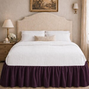 May include: A bed valance in a deep, solid purple colour featuring a gathered, ruffled design. The valance is positioned beneath a white bedspread, with the fabric draping down to the floor, creating a layered appearance for the bed frame.