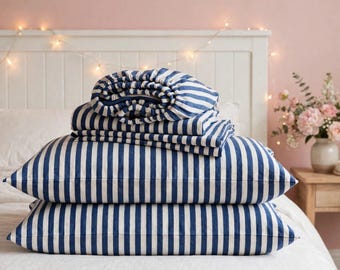 May include: A set of bedding featuring a navy blue and white vertical striped pattern. The stack includes two rectangular pillows and a folded fitted sheet, all made from the same striped fabric, arranged neatly on a white bedspread.