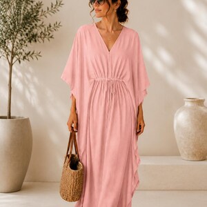 May include: A light pink, V-neck maxi dress with a drawstring waist and flowing sleeves. The dress is worn with sunglasses and a woven tote bag with brown leather handles. The model is barefoot.