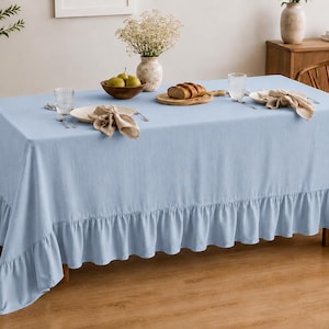 May include: A rectangular light blue tablecloth with a ruffled edge draped over a dining table. The tablecloth features a solid color and a gathered skirt-like ruffle that hangs down from the edges, covering the sides of the table surface.