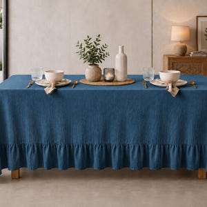 May include: A rectangular blue tablecloth with a ruffled edge draped over a dining table. The table is set with two white bowls, two clear glasses, gold-toned cutlery, a central vase with greenery, a small candle holder, and a tall white bottle.