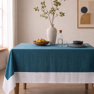 Puede incluir: Un mantel rectangular que presenta un centro de color azul cerceta profundo y un borde ancho de color blanco a lo largo del borde inferior. La tela tiene una textura tejida visible. El mantel cubre una mesa, sirviendo de base para objetos.