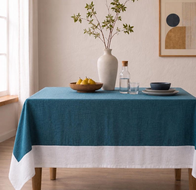 Puede incluir: Un mantel rectangular que presenta un centro de color azul cerceta profundo y un borde ancho de color blanco a lo largo del borde inferior. La tela tiene una textura tejida visible. El mantel cubre una mesa, sirviendo de base para objetos.