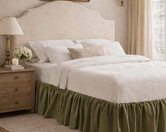 May include: A bedroom scene featuring a bed with a cream-colored headboard and white bedding. The bed has a green bed skirt. A wooden nightstand with a lamp and a painting on the wall complete the scene.
