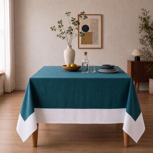 Puede incluir: Un mantel cuadrado que presenta un centro de color verde azulado profundo y un borde blanco ancho en la parte inferior. La tela tiene una textura tejida visible y cae sobre los cuatro lados de una mesa, creando un aspecto geom&eacute;trico bicolor.