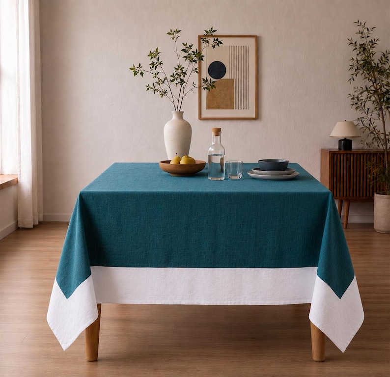 Puede incluir: Un mantel cuadrado que presenta un centro de color verde azulado profundo y un borde blanco ancho en la parte inferior. La tela tiene una textura tejida visible y cae sobre los cuatro lados de una mesa, creando un aspecto geom&eacute;trico bicolor.