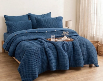 May include: A deep blue waffle-weave duvet cover set featuring a button-down closure on the top edge. The bedding includes two matching rectangular pillow shams and two square pillow shams, all in a textured, grid-like fabric pattern.