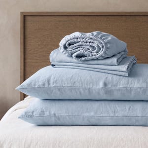 May include: A set of light blue bedding including two rectangular pillowcases stacked horizontally and a folded fitted sheet with elasticated edges resting on top. The fabric has a visible woven texture and a soft, matte finish in a solid colour.