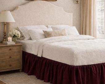 May include: A bedroom scene featuring a bed with a cream-colored headboard and white bedding. The bed has a deep burgundy bed skirt. A wooden nightstand with a lamp and a painting on the wall complete the scene.