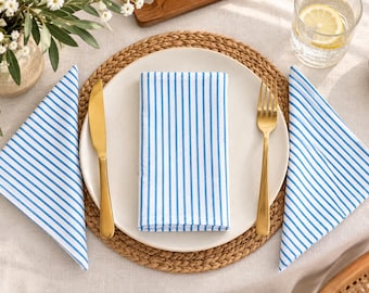 May include: A white dinner plate sits on a round woven placemat, featuring a folded white napkin with thin blue vertical stripes. A gold-toned knife and fork are placed on the plate, flanked by two additional folded napkins with matching blue stripes.