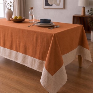 Puede incluir: Un mantel rectangular con un panel central de color naranja quemado s&oacute;lido bordeado por una franja ancha de color beige claro. La tela tiene una textura tejida visible y el mantel cubre una mesa con un cuenco, platos y una jarra de agua.
