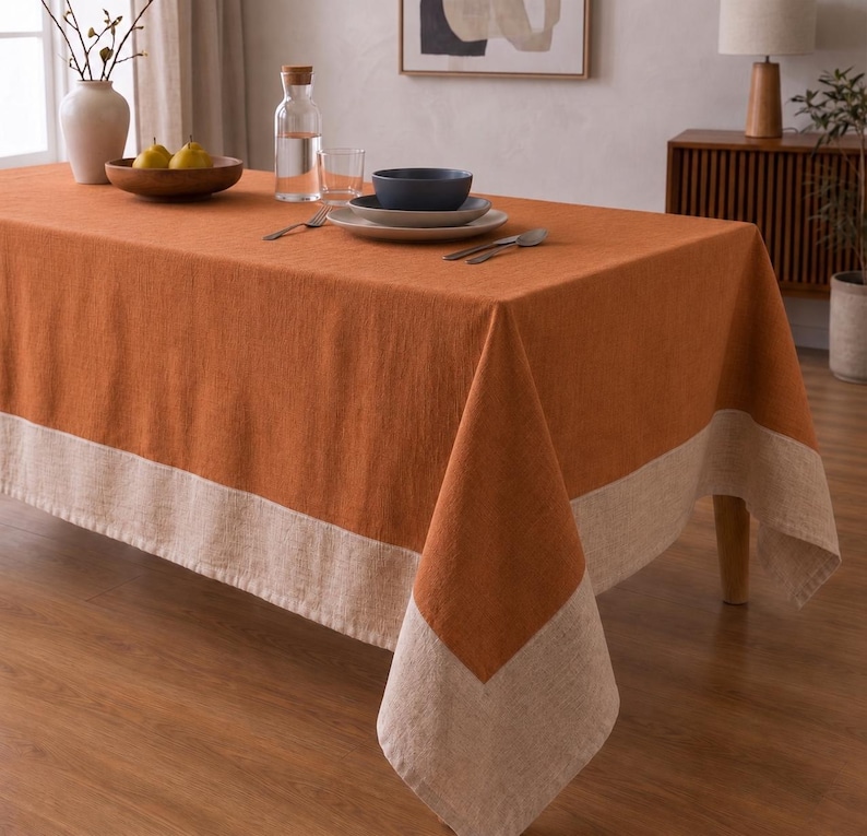 Puede incluir: Un mantel rectangular con un panel central de color naranja quemado s&oacute;lido bordeado por una franja ancha de color beige claro. La tela tiene una textura tejida visible y el mantel cubre una mesa con un cuenco, platos y una jarra de agua.