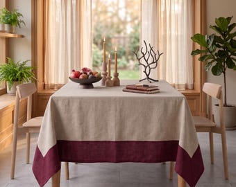 Puede incluir: Una mesa de comedor rectangular cubierta con un mantel de lino beige que presenta un borde ancho de color burdeos oscuro en la parte inferior. La mesa está decorada con un cuenco oscuro con frutas, tres velas altas y dos libros apilados.