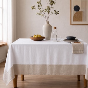 Puede incluir: Un mantel rectangular blanco con un borde ancho de color beige en la parte inferior. La mesa est&aacute; decorada con un gran jarr&oacute;n de cer&aacute;mica color hueso con ramas verdes, un cuenco de madera con peras amarillas, una jarra de cristal y platos.