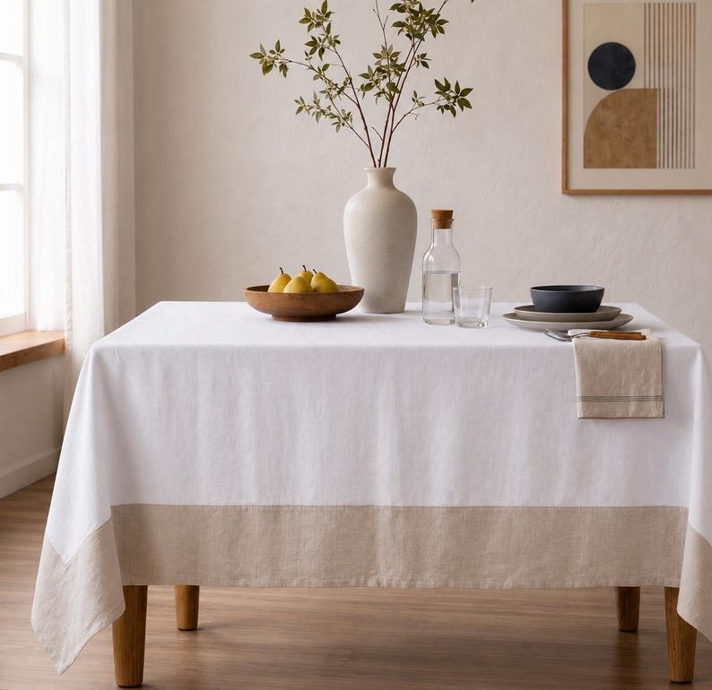 Puede incluir: Un mantel rectangular blanco con un borde ancho de color beige en la parte inferior. La mesa est&aacute; decorada con un gran jarr&oacute;n de cer&aacute;mica color hueso con ramas verdes, un cuenco de madera con peras amarillas, una jarra de cristal y platos.