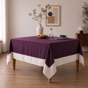 Puede incluir: Un mantel cuadrado con un centro de color morado intenso y un borde ancho en color blanco roto a lo largo del dobladillo inferior. La tela presenta una textura tejida visible que cae sobre las cuatro patas de una mesa de madera.