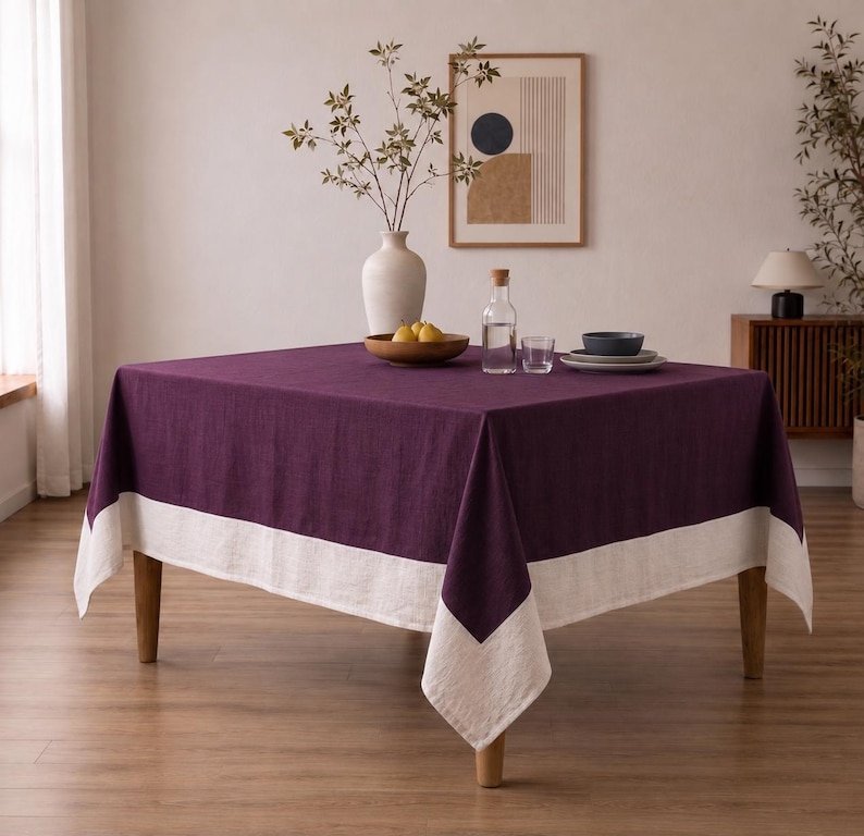 Puede incluir: Un mantel cuadrado con un centro de color morado intenso y un borde ancho en color blanco roto a lo largo del dobladillo inferior. La tela presenta una textura tejida visible que cae sobre las cuatro patas de una mesa de madera.