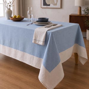 Puede incluir: Un mantel rectangular que presenta un panel central de color azul claro s&oacute;lido con un borde ancho y contrastante en color blanco roto a lo largo de los lados. La tela tiene una textura tejida visible y cae sobre los lados de una mesa de comedor.