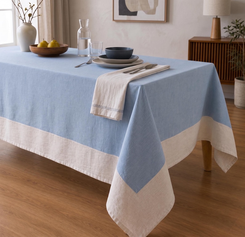 Puede incluir: Un mantel rectangular que presenta un panel central de color azul claro s&oacute;lido con un borde ancho y contrastante en color blanco roto a lo largo de los lados. La tela tiene una textura tejida visible y cae sobre los lados de una mesa de comedor.