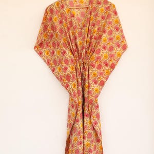 Puede incluir: Un vestido caft&aacute;n con un patr&oacute;n floral repetitivo en tonos rosa y amarillo sobre un fondo crema. La prenda tiene un escote en V, mangas anchas estilo kimono y un detalle de cintura ce&ntilde;ida, colgado de una percha de madera.