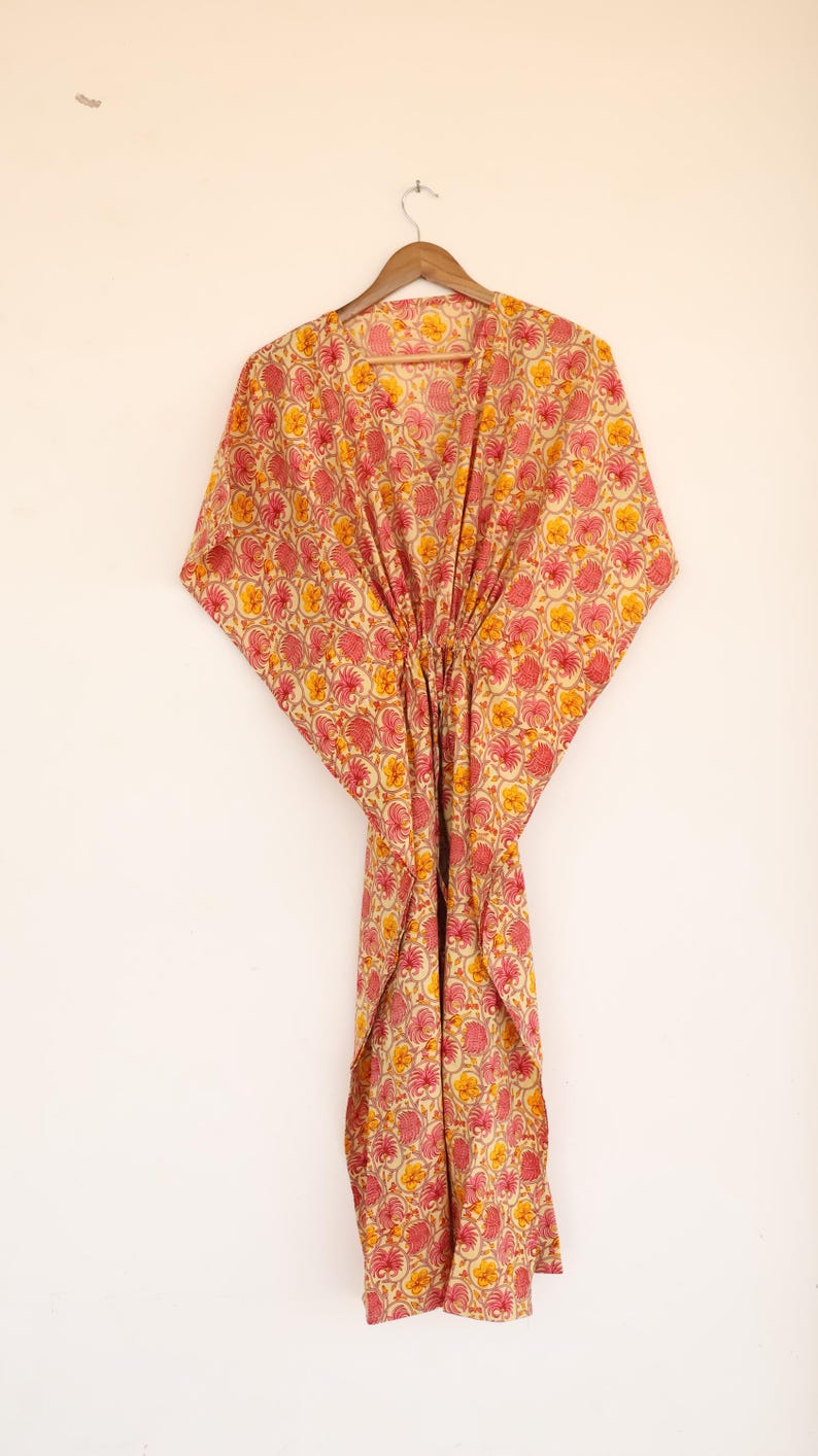 Puede incluir: Un vestido caft&aacute;n con un patr&oacute;n floral repetitivo en tonos rosa y amarillo sobre un fondo crema. La prenda tiene un escote en V, mangas anchas estilo kimono y un detalle de cintura ce&ntilde;ida, colgado de una percha de madera.