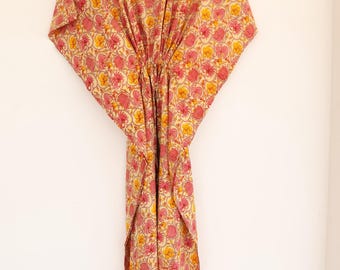 Puede incluir: Un vestido caftán con base de color crema claro y un patrón floral repetitivo en tonos rosa y amarillo. La prenda tiene un escote en V, mangas cortas anchas y un detalle de cintura ceñida, colgado de una percha de madera.