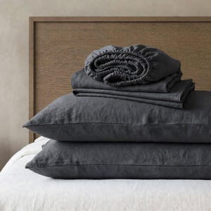 May include: A set of dark charcoal gray bedding including two rectangular pillowcases stacked on top of each other, topped with a folded flat sheet and a fitted sheet with visible elastic edges, all featuring a textured, matte fabric finish.