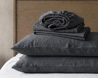 May include: A set of dark charcoal gray bedding including two rectangular pillowcases stacked on top of each other, topped with a folded flat sheet and a fitted sheet with visible elastic edges, all featuring a textured, matte fabric finish.