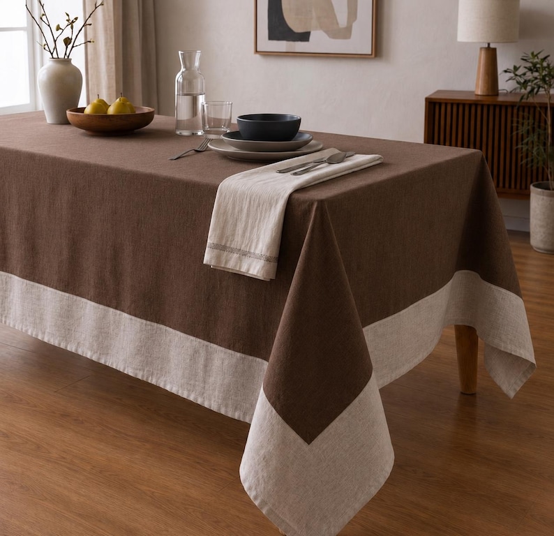 Puede incluir: Un mantel rectangular que presenta un panel central de color marr&oacute;n oscuro s&oacute;lido con un borde ancho y contrastante de color beige claro. La tela tiene una textura tejida visible y el mantel est&aacute; colocado sobre una mesa con vajilla.
