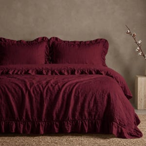 Puede incluir: Un juego de cama de color burdeos intenso que incluye una funda nórdica y dos fundas de almohada a juego, todas decoradas con bordes de volantes. La tela presenta un tejido texturizado visible y un esquema de color rojo oscuro monocromático.