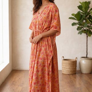 Puede incluir: Un vestido tipo caft&aacute;n largo hasta los pies con un estampado floral en tonos coral y naranja. La prenda presenta un escote en V, mangas cortas holgadas y un cord&oacute;n ajustable en la cintura que crea una silueta fruncida.