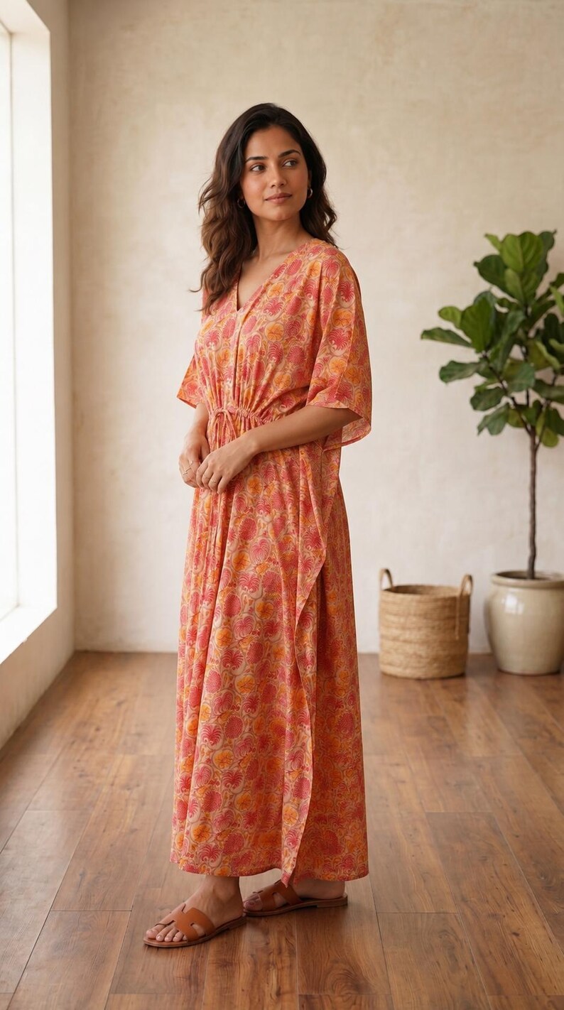 Puede incluir: Un vestido tipo caft&aacute;n largo hasta los pies con un estampado floral en tonos coral y naranja. La prenda presenta un escote en V, mangas cortas holgadas y un cord&oacute;n ajustable en la cintura que crea una silueta fruncida.