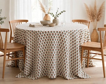 May include: A round dining table covered with a white tablecloth featuring a repeating pattern of small red flowers and green leaves, surrounded by four light brown wooden chairs with vertical back slats and curved armrests.