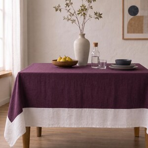 Puede incluir: Un mantel rectangular que presenta una secci&oacute;n principal de color morado ciruela intenso y un borde ancho blanco en contraste a lo largo del borde inferior. La tela tiene una textura tejida visible, drapeada sobre una mesa con aspecto limpio.