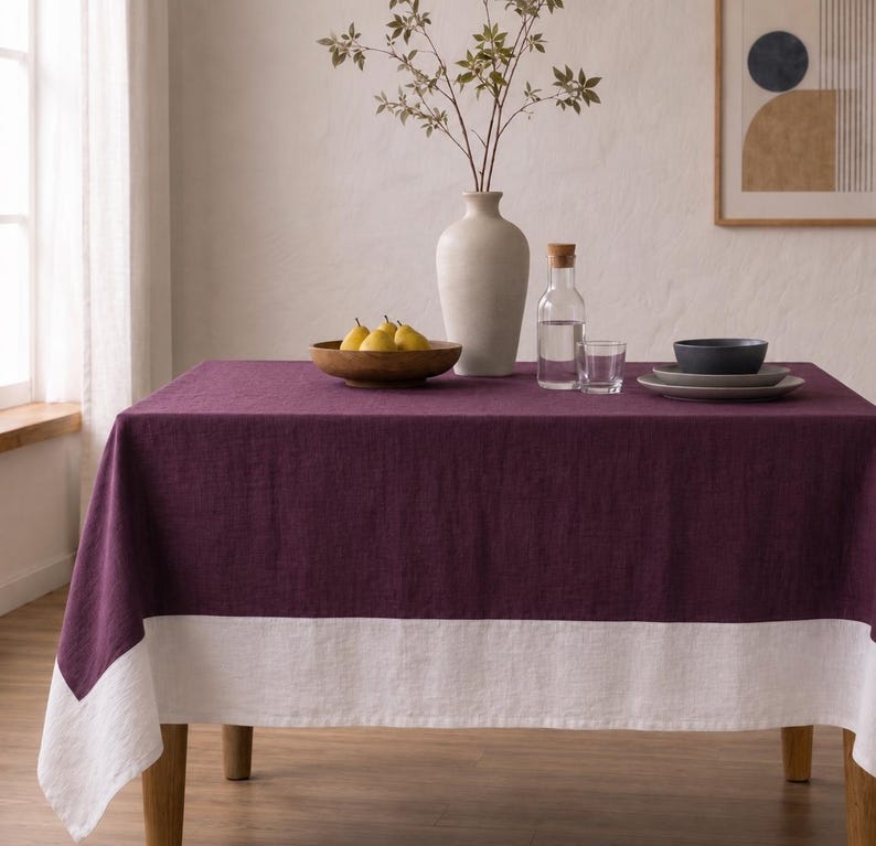 Puede incluir: Un mantel rectangular que presenta una secci&oacute;n principal de color morado ciruela intenso y un borde ancho blanco en contraste a lo largo del borde inferior. La tela tiene una textura tejida visible, drapeada sobre una mesa con aspecto limpio.