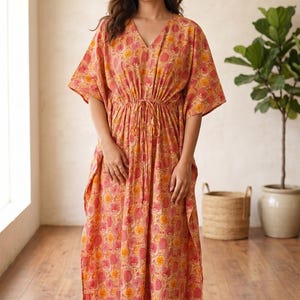 May include: A floor-length kaftan dress featuring a vibrant orange and yellow floral pattern, a V-neckline, short wide sleeves, and a drawstring waist tie that creates a gathered effect. The garment has a loose, flowing silhouette.