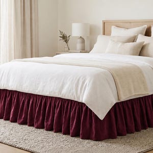 May include: A bed featuring a deep burgundy ruffled bed skirt, a white duvet cover, and a set of beige and white pillows. The bed is positioned against a light wood headboard, creating a layered look with the neutral bedding and dark base.