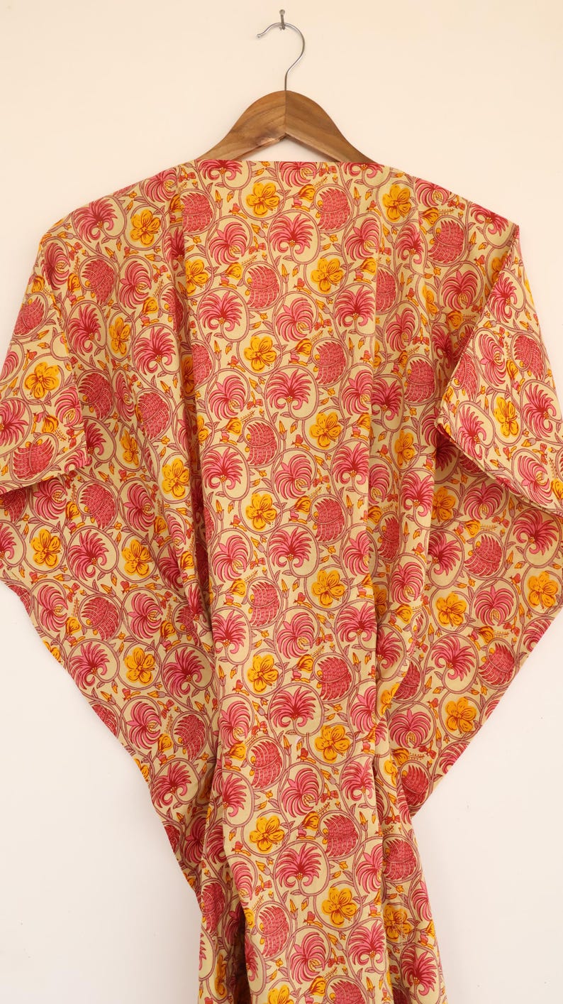Puede incluir: Un caft&aacute;n con base de color crema y un patr&oacute;n repetitivo de follaje rosado similar a palmeras y flores amarillas. La prenda tiene mangas anchas y fluidas y se muestra en una percha de madera sobre un fondo claro y liso.