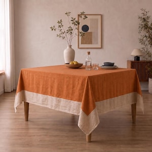 Puede incluir: Un mantel cuadrado que presenta un centro de color naranja quemado vibrante con un borde ancho de color beige natural a lo largo de los lados. La tela tiene una textura tejida visible y cubre una mesa de madera de cuatro patas, creando un aspecto limpio.