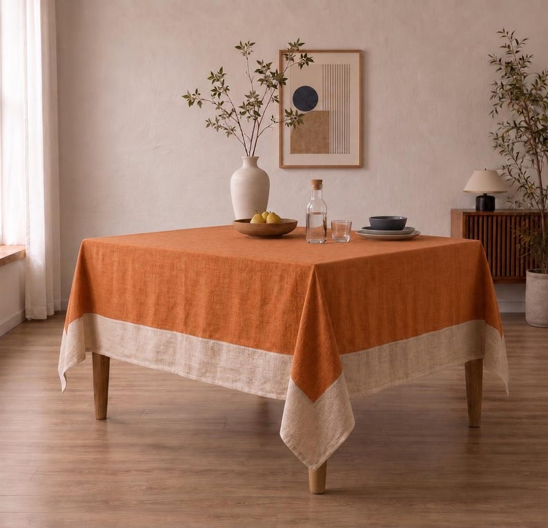 Puede incluir: Un mantel cuadrado que presenta un centro de color naranja quemado vibrante con un borde ancho de color beige natural a lo largo de los lados. La tela tiene una textura tejida visible y cubre una mesa de madera de cuatro patas, creando un aspecto limpio.